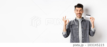 Very good. Smiling guy with plastic credit card showing okay sign, smiling satisfied, recommend bank, standing on white background 103145223