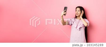 Stylish asian girl blogger taking selfie on mobile phone, posing for smartphone photo, standing in dress against pink background 103145289