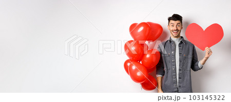 Valentines day and love concept. Cheerful funny guy showing heart cutout, standing near romantic balloons and smiling excited at camera, white background Valentines day and love concept. Cheerful funny guy showing heart cutout, standing near romantic balloons and smiling excited at camera, white background 103145322
