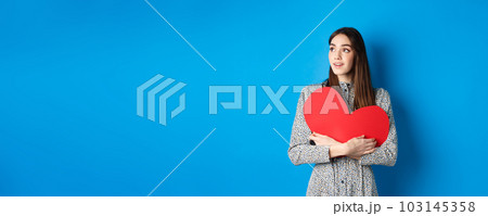 Valentines day. Hopeful girl dreaming off soulmate, looking aside at empty space with excitement, hugging big red heart cutout, standing on blue background Valentines day. Hopeful girl dreaming off soulmate, looking aside at empty space with excitement, hugging big red heart cutout, standing on blue background 103145358