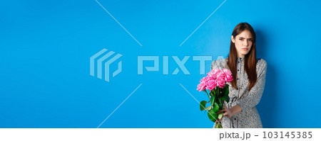 Valentines day concept. Displeased young woman rejecting flowers gift, frowning and grimacing, refusing take roses, standing on blue background 103145385
