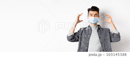 Health, covid and pandemic concept. Funny guy put on medical mask and look at camera, prevent catching coronavirus, standing on white background 103145386