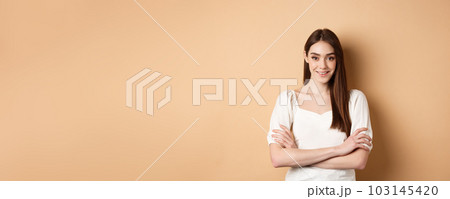 Smiling candid girl in dress cross arms on chest, looking like professional, standing confident on beige background Smiling candid girl in dress cross arms on chest, looking like professional, standing confident on beige background 103145420