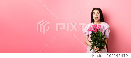 Korean teen girl in dress having romantic date on Valentines day, holding bouquet of roses and looking surprised at camera, receive gift on date from lover, pink background 103145459
