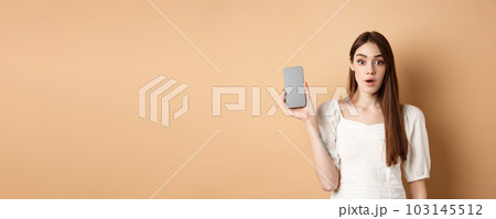 Excited pretty girl showing empty cell phone screen, look amazed at camera, standing on beige background 103145512