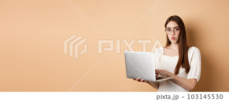 Concerned girl in glasses look at camera confused, working on laptop, having trouble with computer, standing on beige background 103145530