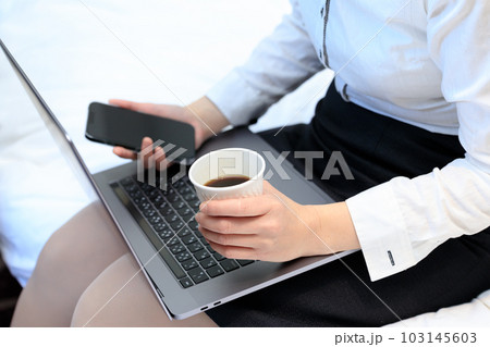 ベッドに座ってコーヒーを飲みながらスマホとパソコンを使用している女性 103145603