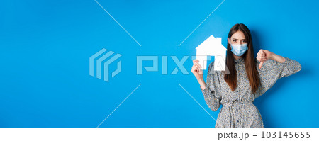 Real estate, covid-19 and pandemic concept. Upset female client in medical mask showing paper house cutout and thumbs down, frowning disappointed, blue background 103145655