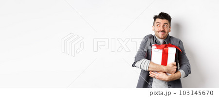 Happy boyfriend hugging gift from lover, looking aside at empty space and smiling excited, making romantic present on valentines day, standing on white background 103145670
