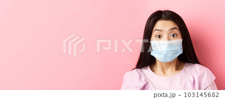 Covid-19 and healthy people concept. Close-up portrait of surprised asian girl in medical mask looking excited at camera, standing against pink background 103145682