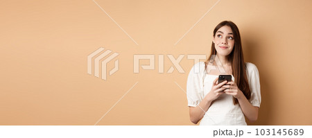 Romantic girl thinking with phone, looking aside and smiling dreamy, using dating app on smartphone, standing on beige background 103145689