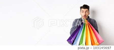 Excited man holding colorful shopping bags and looking left at special promo offer in store, standing on white background 103145697