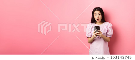 Online shopping. Surprised asian woman in cute dress, open mouth amazed after reading news on smartphone, standing on pink background 103145749