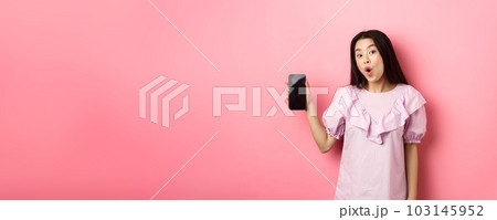 Online shopping. Excited asian woman say wow, showing empty smartphone screen and looking amazed at camera, standing against pink background Online shopping. Excited asian woman say wow, showing empty smartphone screen and looking amazed at camera, standing against pink background 103145952