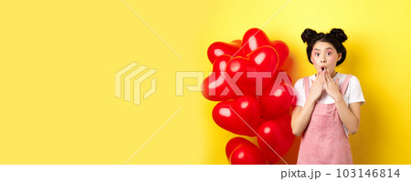 Valentines day concept. Impressed korean girl say wow, gasping and holding hands near opened mouth, staring at camera surprised, standing near romantic red heart balloons, yellow background 103146814