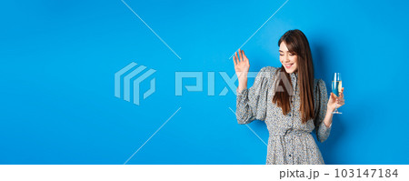 Celebration and party concept. Dancing girl holding glass of champagne, having fun at holiday, celebrating birthday, standing on blue background 103147184