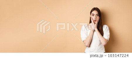 Wow unbelievable. Impressed young woman gasping and looking astonished by news, staring at camera, touching face in awe, standing on beige background 103147224