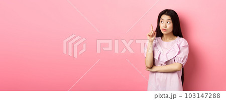 Beautiful asian girl raising finger and looking left at logo, having an idea, found solution, standing against pink background Beautiful asian girl raising finger and looking left at logo, having an idea, found solution, standing against pink background 103147288