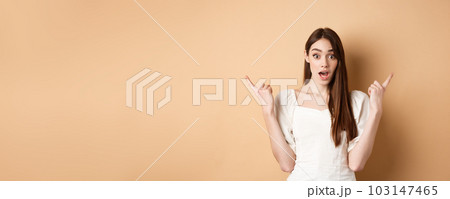 Surprised young girl pointing sideways at left and right, gasping and saying wow amazed, showing two cool choices, beige background 103147465