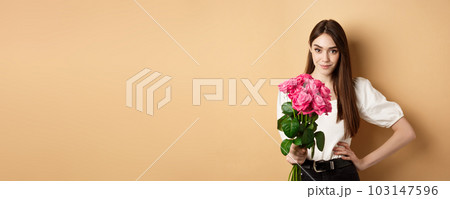 Valentines day. Beautiful girlfriend holding pink roses and looking at camera. Young woman receive flowers from her date, standing on beige background Valentines day. Beautiful girlfriend holding pink roses and looking at camera. Young woman receive flowers from her date, standing on beige background 103147596
