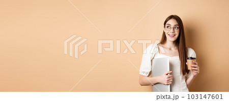 Happy young woman drinking coffee and holding laptop, going study, looking aside with cheerful smile, standing on beige background 103147601
