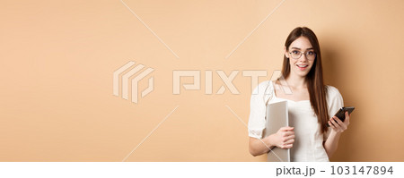 Smiling young woman in glasses reading exciting news on mobile phone, holding laptop and looking happy at camera, standing on beige background Smiling young woman in glasses reading exciting news on mobile phone, holding laptop and looking happy at camera, standing on beige background 103147894