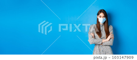 Covid-19, pandemic lifestyle concept. Serious young woman with long natural hair, wearing medical mask, cross arms on chest, protect herself from virus, blue background Covid-19, pandemic lifestyle concept. Serious young woman with long natural hair, wearing medical mask, cross arms on chest, protect herself from virus, blue background 103147909