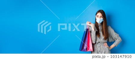 Covid-19, pandemic and lifestyle concept. Girl wearing medical mask on shopping in malls, holding paper bags and look aside at empty space, standing on blue background 103147910