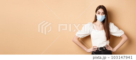 Covid-19 and lifestyle concept. Woman in medical mask looking alarmed at camera, standing on beige background 103147941