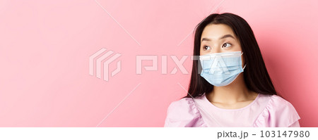 Covid-19, pandemic and quarantine concept. Close-up of dreamy asian girl in medical mask looking left at logo with thoughtful face, standing on pink background Covid-19, pandemic and quarantine concept. Close-up of dreamy asian girl in medical mask looking left at logo with thoughtful face, standing on pink background 103147980