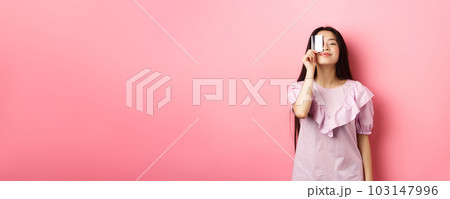 Shopping. Cute candid woman smiling, showing plastic credit card on face, standing in dress against pink background 103147996