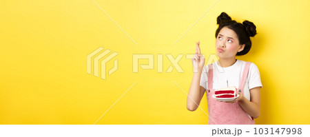 Holidays and celebration. Cute asian birthday girl making wish at party wish cake and candle, cross fingers and looking up, standing on yellow background 103147998