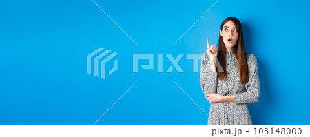 Thoughtful attractive girl in dress pitching an idea, raising finger and looking up, have a plan, standing on blue background 103148000