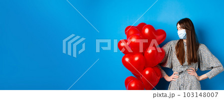 Covid-19 and Valentines day. Young bothered girl in medical mask and dress, looking at heart balloons, waiting on date, standing blue background 103148007