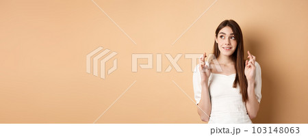 Hopeful cute woman looking at upper left corner space and cross fingers for good luck, making wish, praying for dream to come true, standing on beige background Hopeful cute woman looking at upper left corner space and cross fingers for good luck, making wish, praying for dream to come true, standing on beige background 103148063