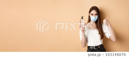 Covid-19 and preventive measures concept. Smiling girl in medical mask show thumb up and hand sanitizer, recommend product for disinfection, beige background Covid-19 and preventive measures concept. Smiling girl in medical mask show thumb up and hand sanitizer, recommend product for disinfection, beige background 103148070