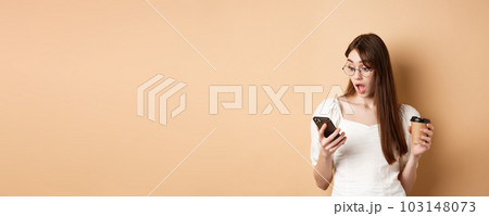 Surprised girl reading exciting news on cell phone, drop jaw amazed, standing with coffee cup on beige background Surprised girl reading exciting news on cell phone, drop jaw amazed, standing with coffee cup on beige background 103148073