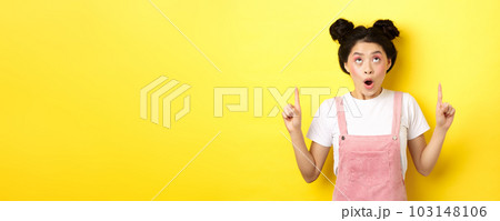 Excited asian teen student with glamour makeup, gasping amazed pointing fingers up, showing advertisement, yellow background 103148106