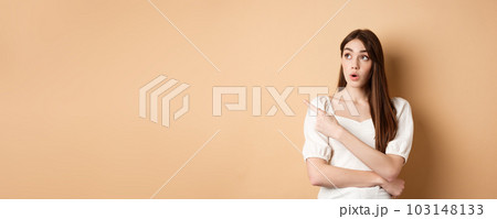 Young woman in dress looking and pointing at upper left corner wondered, saying wow, checking out advertisement with intrigued face, beige background Young woman in dress looking and pointing at upper left corner wondered, saying wow, checking out advertisement with intrigued face, beige background 103148133