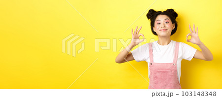 Happy teen koren girl with beauty makeup, showing okay signs and smiling satisfied, recommending product, praise good choice, standing on yellow background 103148348