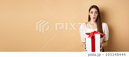 Image of surprised girl receive gift on Valentines day, looking with disbelief and holding present, standing on beige background Image of surprised girl receive gift on Valentines day, looking with disbelief and holding present, standing on beige background 103148444