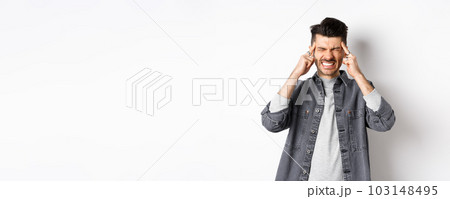 Man with headache grimacing and clenching teeth from painful migraine, touching head temples, suffering migraine, need painkillers, standing on white background 103148495