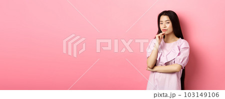 Hmm, interesting. Pensive and cunning asian girl having idea, plan to do something, looking at camera and touching chin with thoughtful face, standing on pink background 103149106