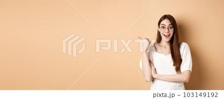 Excited girl in glasses pointing aside at logo, smiling amazed, recommending company, standing on beige background 103149192