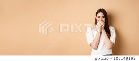 Happy girl laughing and covering mouth with hands, hiding her smile and chuckle over something funny, standing on beige background 103149195