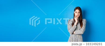 Sensual woman in dress biting finger and looking pensive at camera, standing dreamy against blue background 103149201