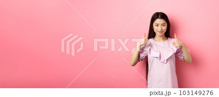 Well done. Smiling asian girl in dress showing thumbs up, like and approve product, praise great deal, standing against pink background 103149276