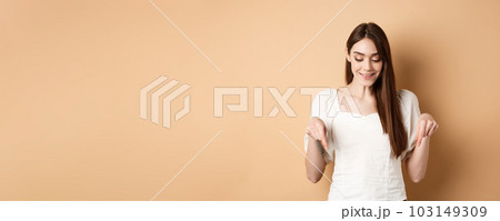 Smiling beautiful female model looking and pointing down with pleased happy face, showing something romantic, standing on beige background 103149309