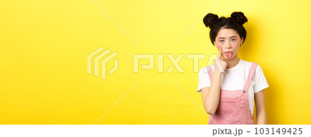 Rude asian girl stretch eyelid and showing tongue, mocking someone, standing on yellow background 103149425