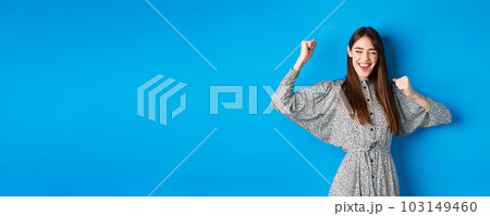 Excited smiling girl showing tongue and dancing with raised hands, celebrating victory or achievement, winning prize, standing happy on blue background 103149460